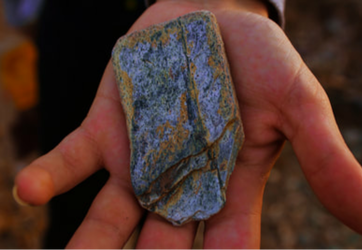 A hand holding a large flat rock with two lines forming an uneven cross. The stone is yellow along the edges and lines, with the rest being patches of white, green, and brown.